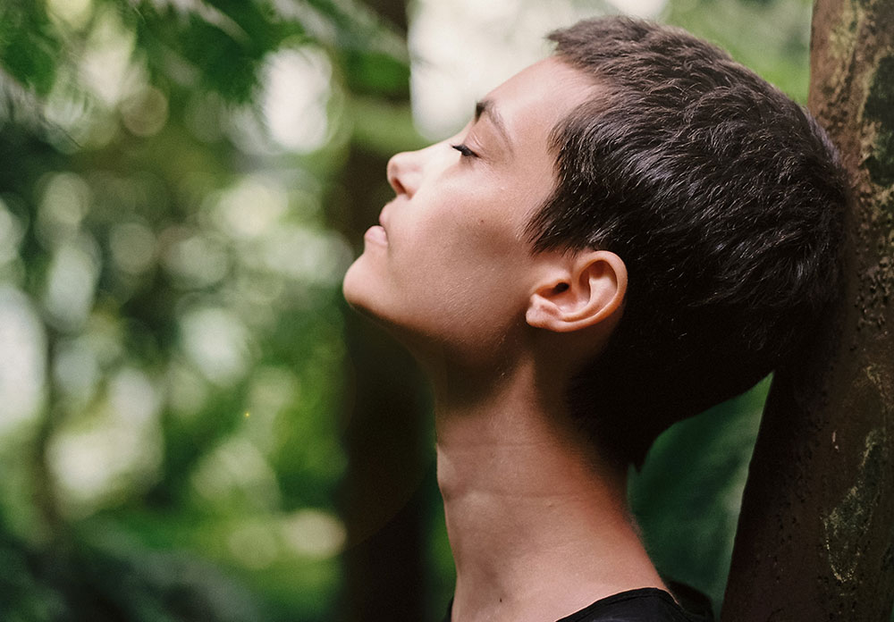 A woman with her eyes close is resting her head against a tree trunk.