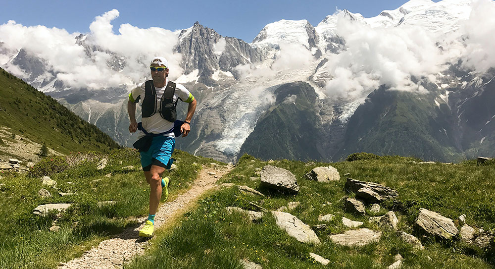 A man wearing sportswear and sunglasses is running along a path in the mountains.