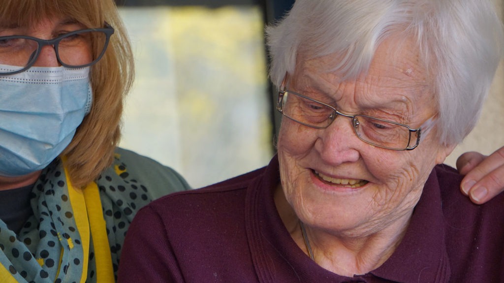 An older woman wearing a face mask has her arm on the shoulder of an elderly woman.