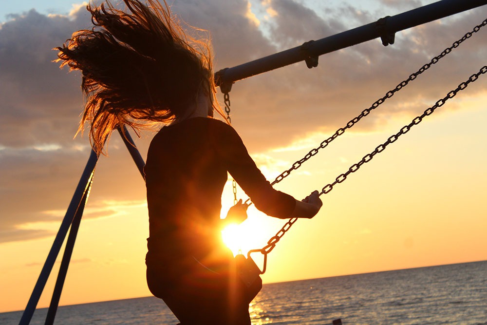 A woman is swinging high on a swing at the beach. The sun is setting in front of them.