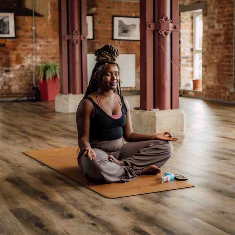 A Woman sitting comfortably cross legged in a yoga studio. ,A POWERbreathe Plus and Smart Adaptor and phone are on the floor next to the mat.