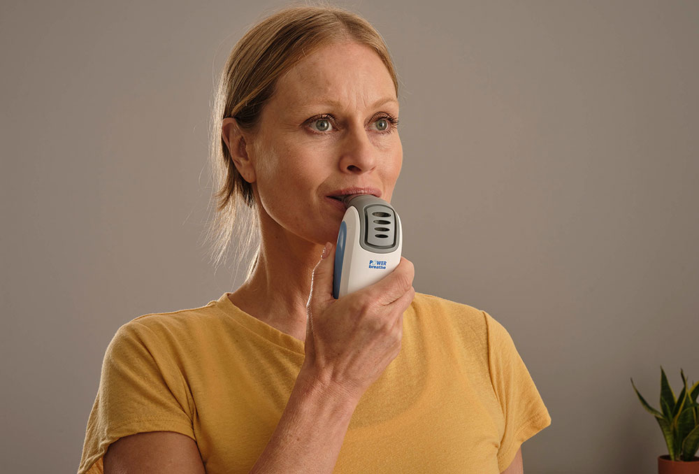 A woman wearing a yellow t-shirt is using a POWERbreathe IMT device.