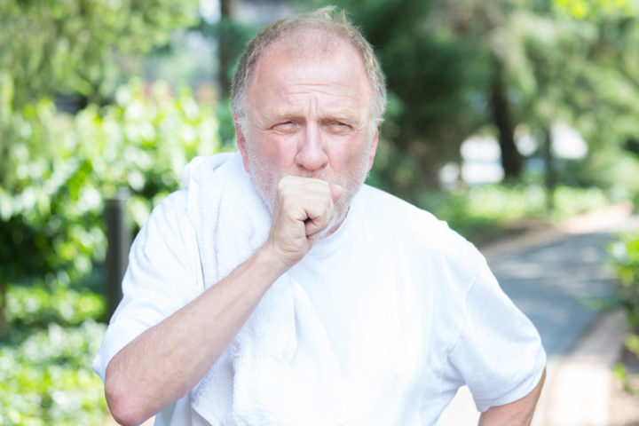 An older man is running outside, he is covering his mouth with his hand which is in a fist.