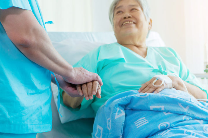 A lady sat in a hospital bed is holding a person's hand - she is smiling up at them.