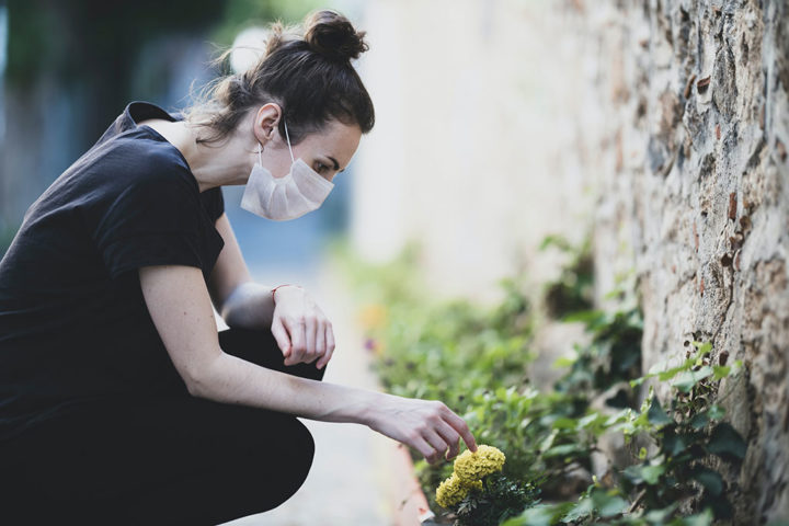 A woman wearing a face mask is crouched down at a flower bed. They are pointing at a yellow flower.
