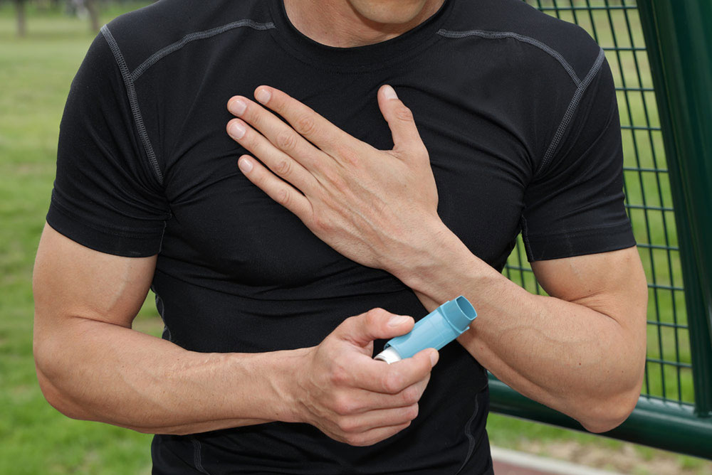 A man in sportswear is holding his chest with one hand, and is holding an inhaler in the other hand.