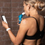 An over the shoulder view of a sporty woman using a Smart Adaptor with a Blue POWERbreathe Plus, looking at the App results on a phone.