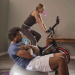 A man leaning back on a balance ball with a Blue Plus in his mouth and a woman on a Wattbike with a Red Plus in her mouth.