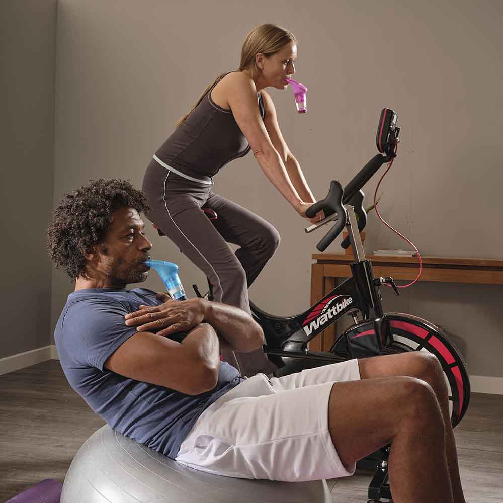 A man leaning back on a balance ball with a Blue Plus in his mouth and a woman on a Wattbike with a Red Plus in her mouth.