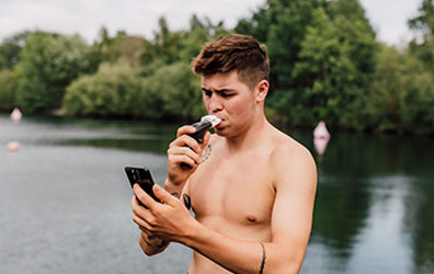Triathlete in wetsuit, stood in front of a lake, training with the POWERbreathe Plus and Smart Adaptor while viewing ActiBreathe app on mobile.