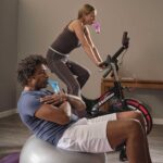 A man leaning back on a balance ball with a Blue Plus in his mouth and a woman on a Wattbike with a Red Plus in her mouth.